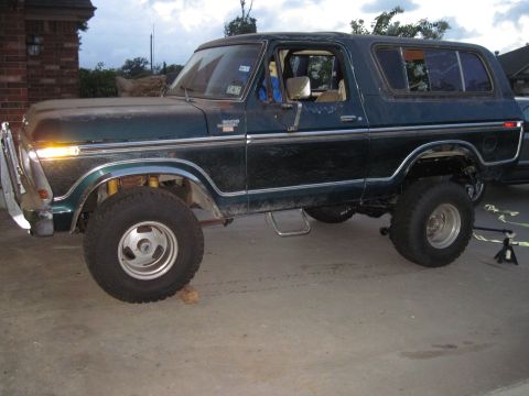 1979 Ford Bronco - A dark green 1979 Ford Bronco is parked on a concrete surface, with its front wheel elevated on a wooden block and a jack stand supporting…