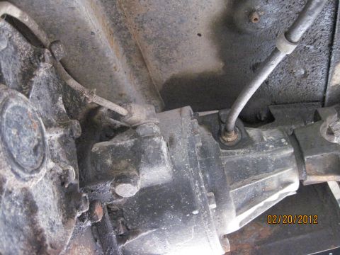 1979 Ford Bronco - A close-up, low-angle shot shows the underside of a vehicle's drivetrain, focusing on the transmission and transfer case area. A metal brak…