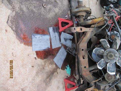 1979 Ford Bronco - A disassembled vehicle front end, likely a 1979 Ford Bronco, lies on a concrete surface with oil stains and absorbent pads. Visible parts i…