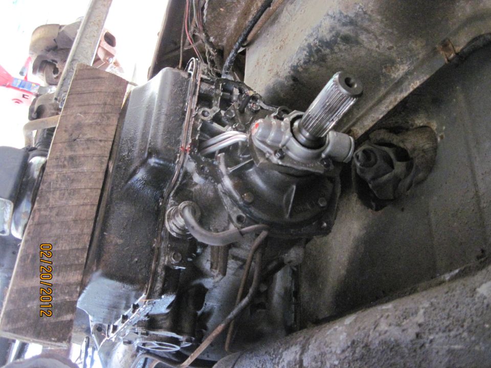 Underside view of a vehicle's transmission and driveshaft with wooden support and visible date stamp
