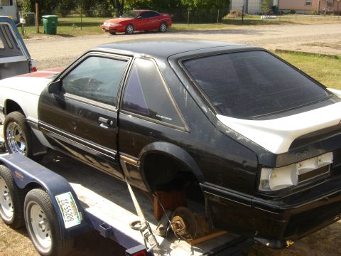 1987 Ford Mustang - A black and white 1987 Ford Mustang coupe is seen on a trailer, missing its rear wheels and with some body damage.