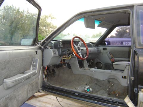 1987 Ford Mustang - The interior of a 1987 Ford Mustang shows a disassembled dashboard and exposed wiring, with a wooden steering wheel.