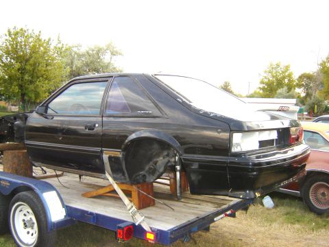 1987 Ford Mustang - A black 1987 Ford Mustang coupe is seen on a trailer, missing its wheels and undergoing some form of work or transport.