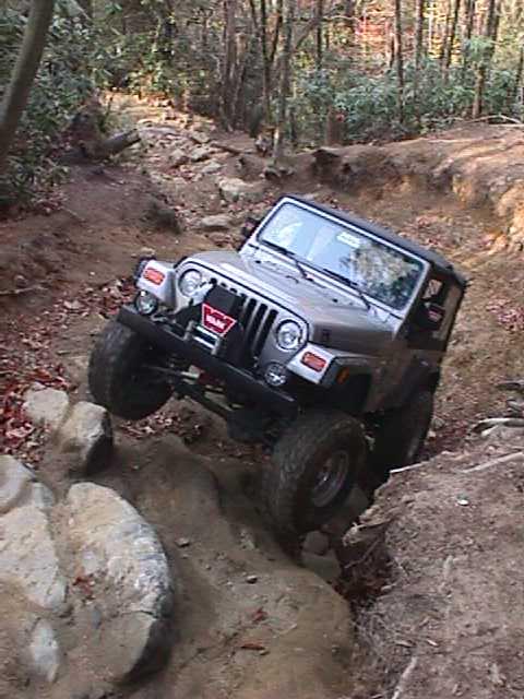 1996 Ford Bronco - A silver Jeep Wrangler navigates a challenging off-road course through a forest.