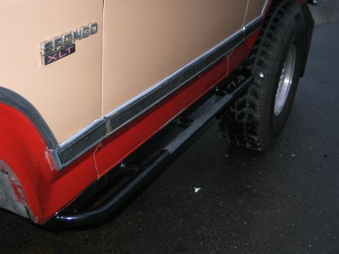 1984 Ford Bronco - Side view of a tan and red 1984 Ford Bronco XLT, equipped with black side steps and large off-road tires.