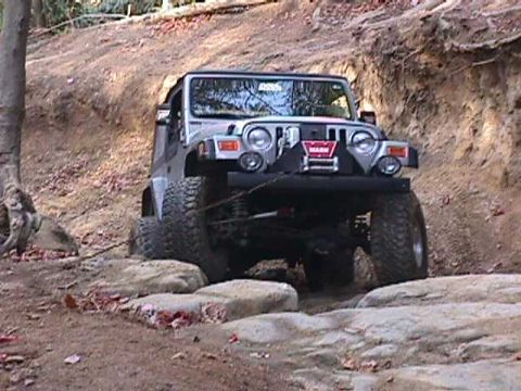 1996 Ford Bronco - A Jeep Wrangler is shown tackling a rocky off-road course with steep dirt embankments.