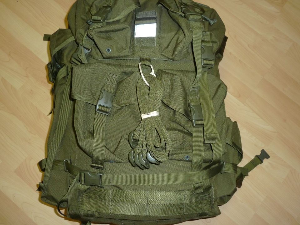 Olive green backpack with multiple straps and buckles, a bundle of green cord tied with white elastic, and a reflective patch on top