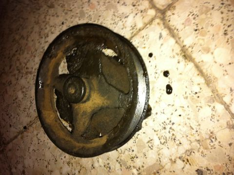1998 Ford Crown Victoria - A greasy power steering pump pulley lies on a speckled tile floor with several oil droplets nearby.