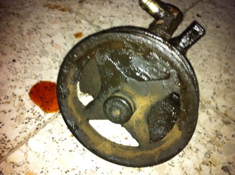 1998 Ford Crown Victoria - A greasy power steering pump sits on a tiled floor next to a small puddle of red fluid.