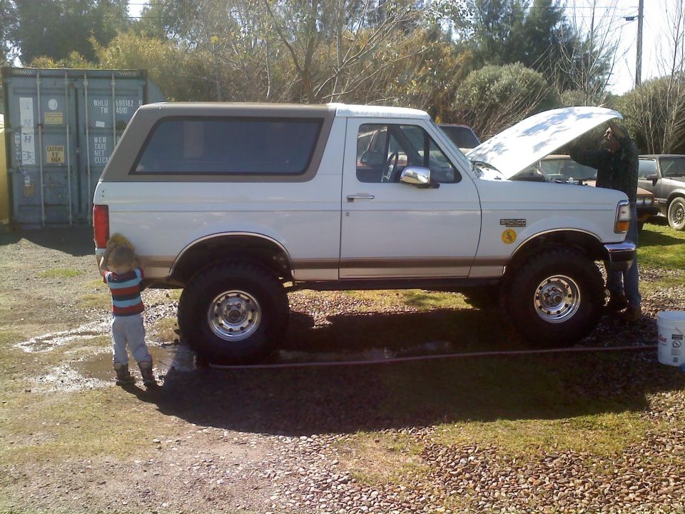 White Ford Bronco, lifted with big tires, hood open, someone working on engine. Kid by rear wheel