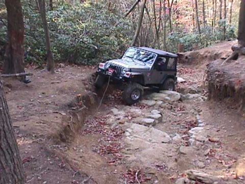 1996 Ford Bronco - A gray Jeep Wrangler navigates a challenging rocky path in a wooded area.