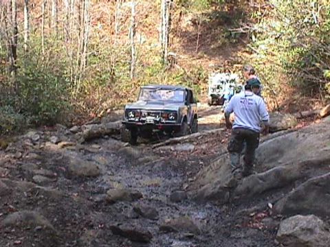 1996 Ford Bronco - Two off-road vehicles traverse a challenging rocky path in a wooded area.