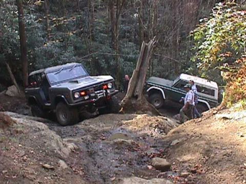1996 Ford Bronco - Two Ford Broncos navigate a challenging off-road course in a wooded area.