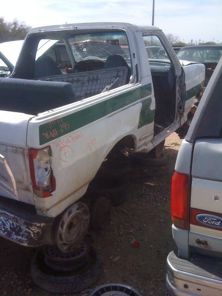 1979 Ford Bronco - A white 1979 Ford Bronco with a green stripe sits in a junkyard, stripped of its doors and wheels.
