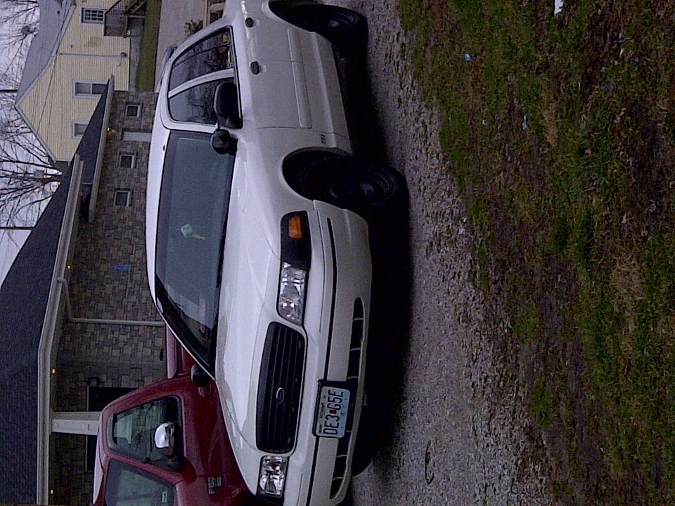 White 2000 Ford Crown Victoria parked on gravel, front 3/4 view, with a red pickup truck to its left