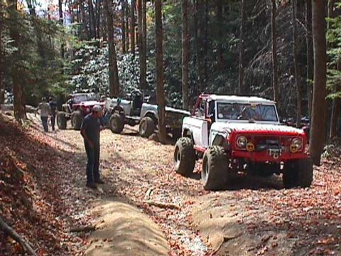 1996 Ford Bronco - A modified red and white off-road vehicle navigates a wooded trail with other vehicles in the background.