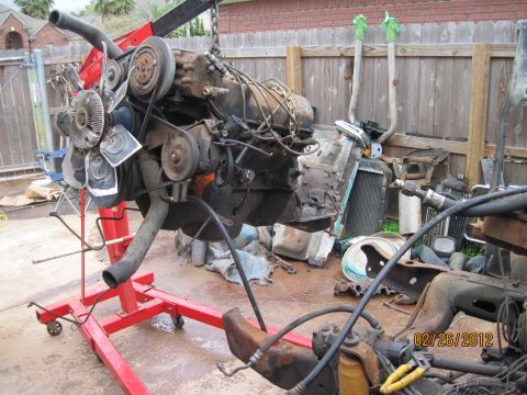 1979 Ford Bronco - An engine is hoisted on a red stand, surrounded by various automotive parts and debris in an outdoor setting.