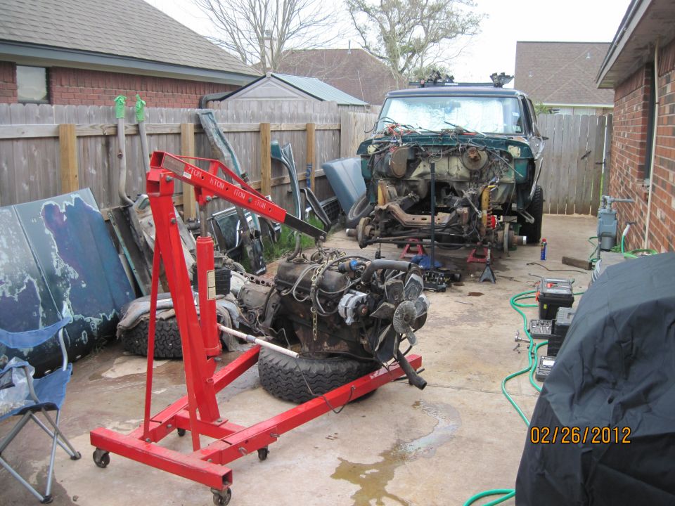Green 1978 Ford Bronco with front end disassembled, engine removed, on jack stands in a backyard