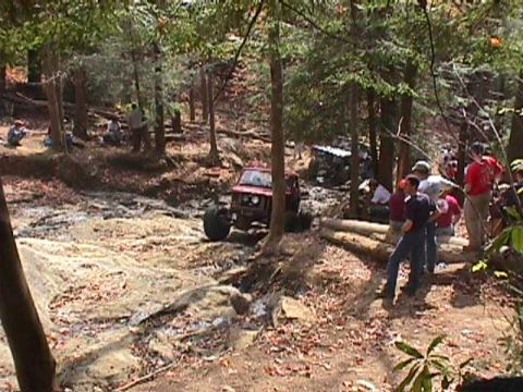 1996 Ford Bronco - A group of people watch as an off-road vehicle makes its way through a challenging, muddy terrain in a forest.