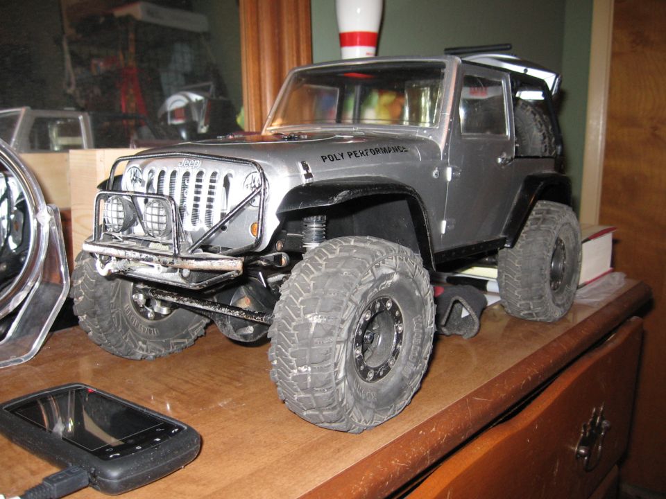 Silver remote-controlled Jeep Wrangler toy with "POLY PERFORMANCE" text on fender, sits on a wooden surface with a black electronic device in foreground