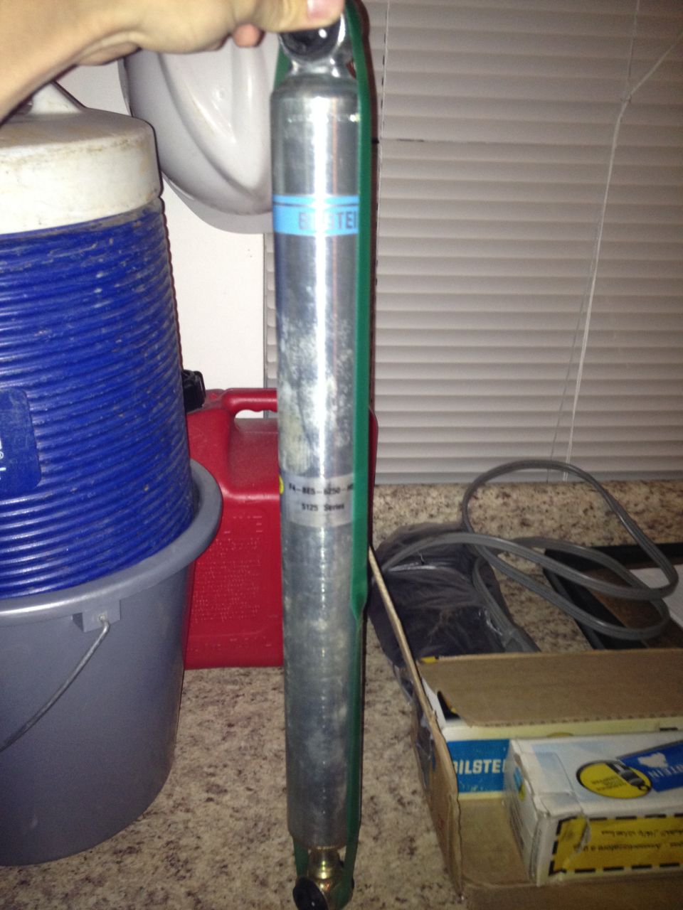 Bilstein shock absorber with blue and green labeling, held by a hand. A red gas can and blue cooler are in the background