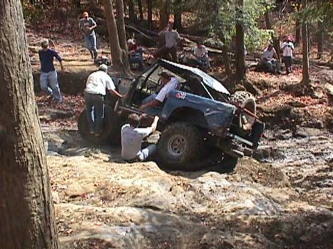 1996 Ford Bronco - A group of people are gathered around a blue 1996 Ford Bronco as it navigates a challenging off-road course.