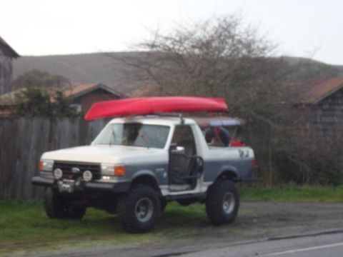 1991 Ford Bronco - A two-tone white and grey Ford Bronco is parked outdoors with a red canoe secured to its roof rack.
