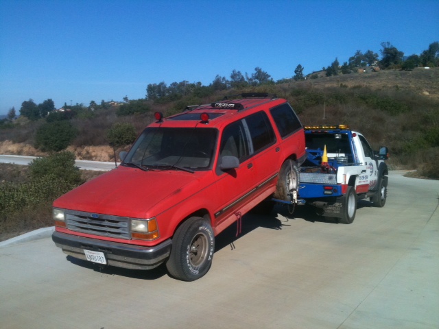 Red Ford SUV lifted at the rear by a white tow truck on a rural concrete road
