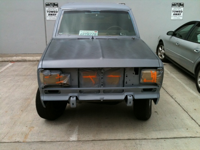 Front view of an SUV, unpainted, with orange tape on headlights and grille area. Another car is parked to the right