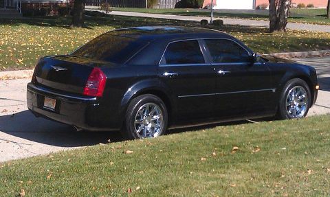 1999 Mercury Grand Marquis - Rear three-quarter view of a black Chrysler 300 sedan on chrome wheels parked along a leaf-strewn residential curb.