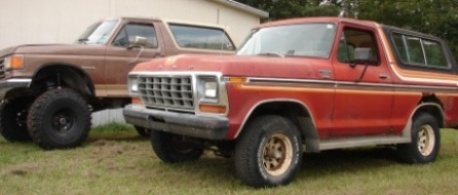 1978 Ford Bronco - Two Ford Broncos, one red and one brown, are parked on a grassy area in front of a building.