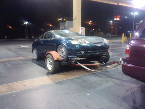 2003 Hyundai Tiburon - A dark blue coupe is loaded onto a trailer, hitched to a maroon SUV, at a gas station after dark.