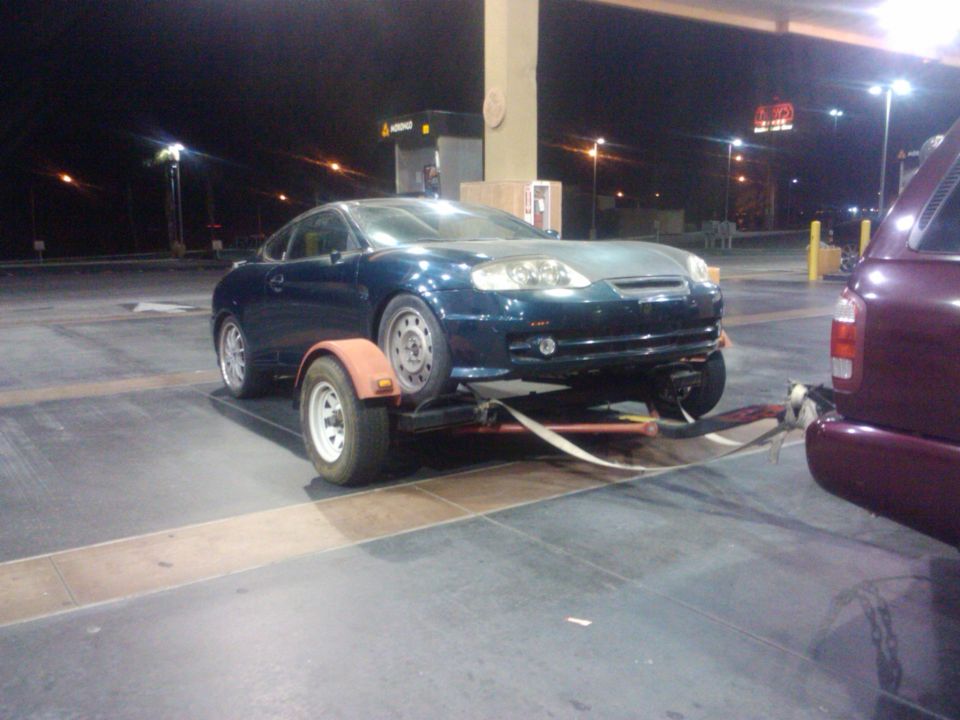 Dark blue coupe on a trailer being towed by a maroon SUV at a gas station at night