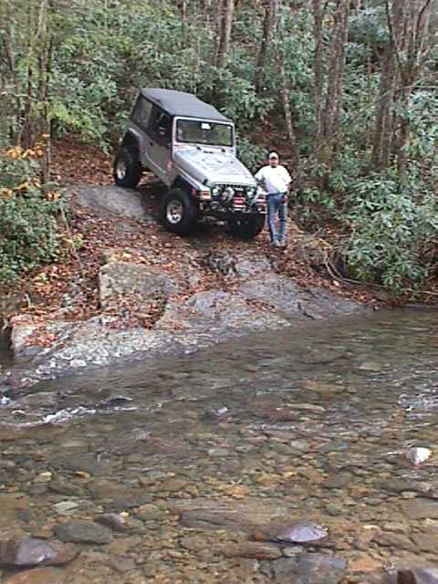 1996 Ford Bronco - A man stands beside a silver Jeep Wrangler as it navigates a rocky, wooded trail next to a shallow stream.