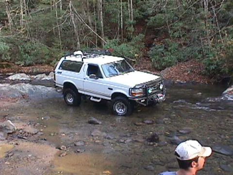 1996 Ford Bronco - A white Ford Bronco navigates a rocky stream bed surrounded by dense forest.