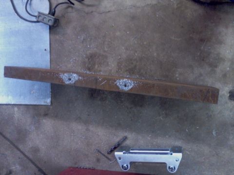 1989 Ford Bronco - A metal bracket with holes drilled into it, likely for mounting purposes, rests on a workshop floor.