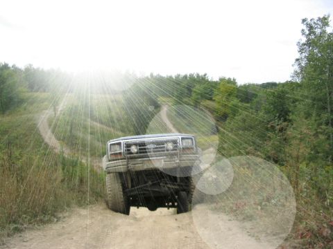 1986 Ford Bronco - An SUV travels down a dusty, unpaved road surrounded by dense foliage under a bright sky.