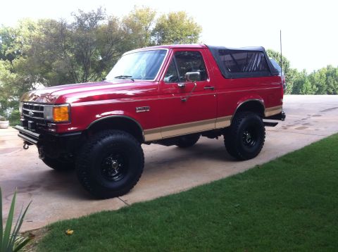 1989 Ford Bronco - A red 1987 Ford Bronco with a tan stripe and black soft top is shown from a three-quarter front angle.