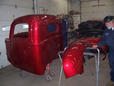 1978 Ford F-150 - A man works on a shiny red pickup truck cab and fender in a garage, with another vehicle visible in the background.