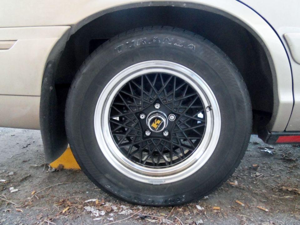 Close-up of a black mesh wheel with a chrome lip on a tan car. Tire has 'TIRANZA' visible