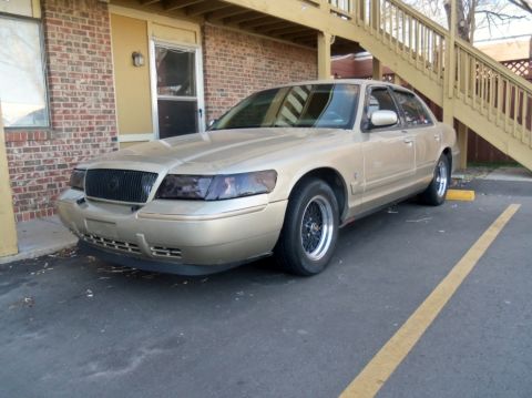 2000 Mercury Grand Marquis - A tan 2000 Mercury Grand Marquis sedan is parked in front of a brick building with a wooden staircase.