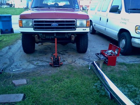 1989 Ford Bronco - A red Ford truck is jacked up in front of a white van, with a bumper and toolbox nearby.