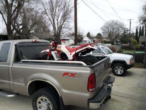 1994 Jeep Cherokee - A white Jeep Cherokee is parked beside a pickup truck that is transporting a red and white dirt bike.