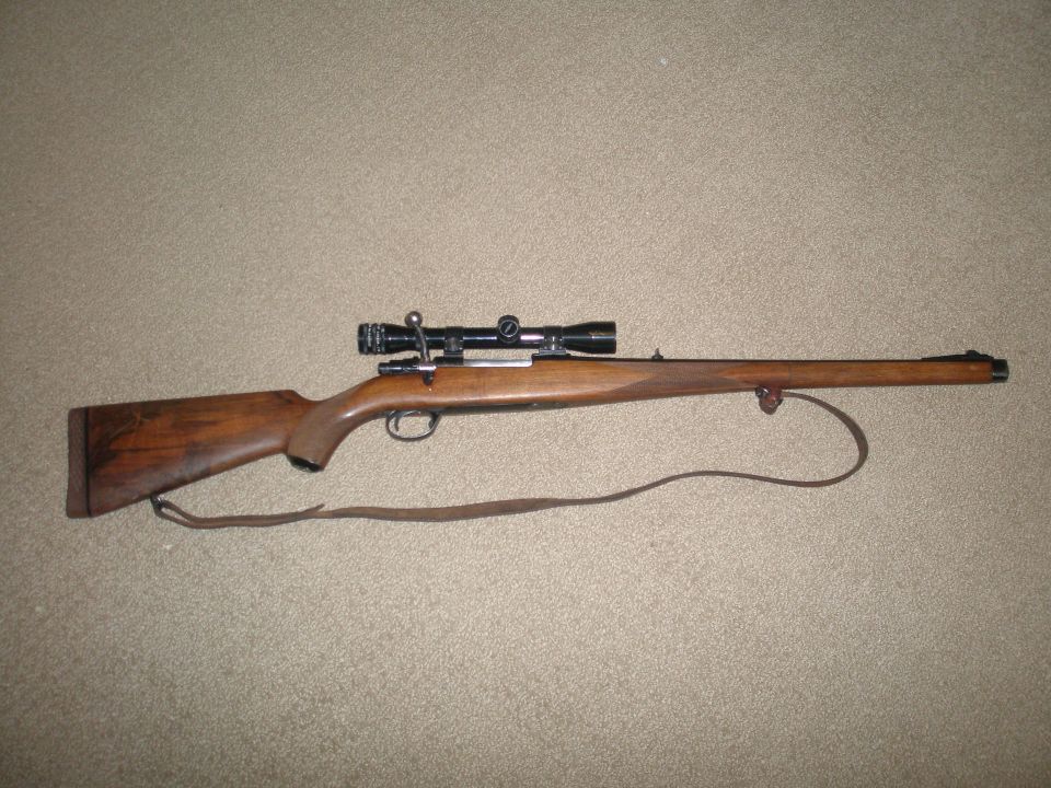 Bolt-action rifle with a scope mounted on top, resting on a carpeted surface