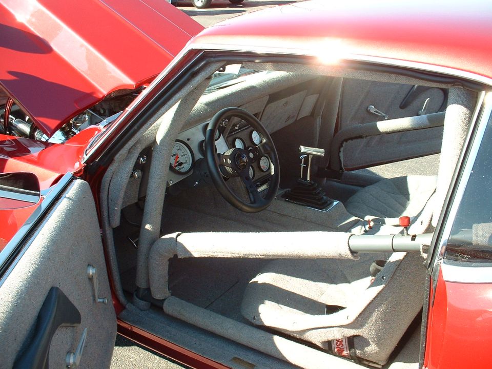 Interior of a red race car with roll cage, racing seats, and aftermarket steering wheel. Sun glare is visible