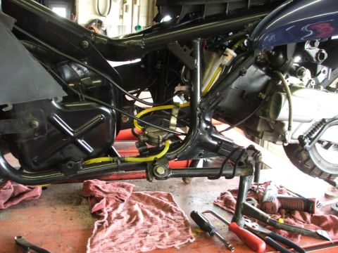 2008 Yamaha Zuma - Close-up of a scooter's internal frame, engine components, and fuel lines, with tools scattered on the workshop floor.