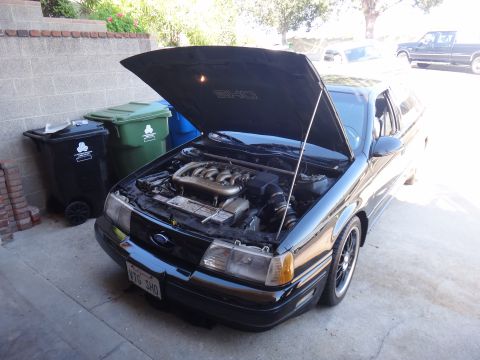 1993 Ford Taurus - The engine bay of a black 1993 Ford Taurus SHO is visible with the hood raised, parked outdoors next to trash bins.