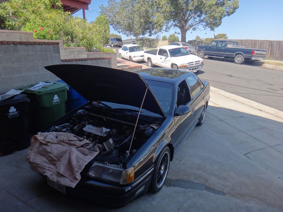 Black sedan with hood open, parked on driveway. Other vehicles visible in background