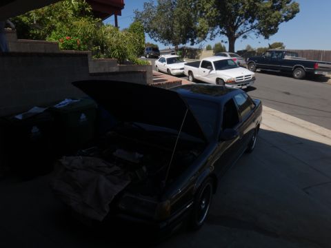 1993 Ford Taurus - A black sedan sits in a driveway with its hood open, alongside a white pickup truck and a dark pickup truck.
