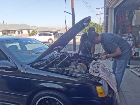 1993 Ford Taurus - Two individuals are gathered around the open engine bay of a black sedan, appearing to perform maintenance.